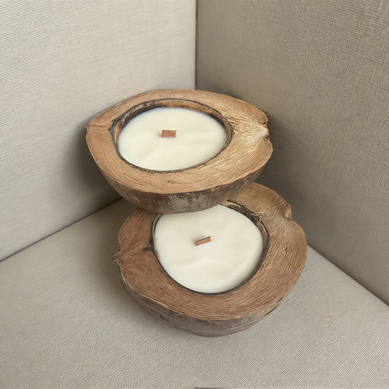 May include: Two coconut shell candles with creamy white wax and wooden wicks. The candles are nestled on a neutral-toned surface, creating a natural and relaxing aesthetic. These candles are a unique home decor item.