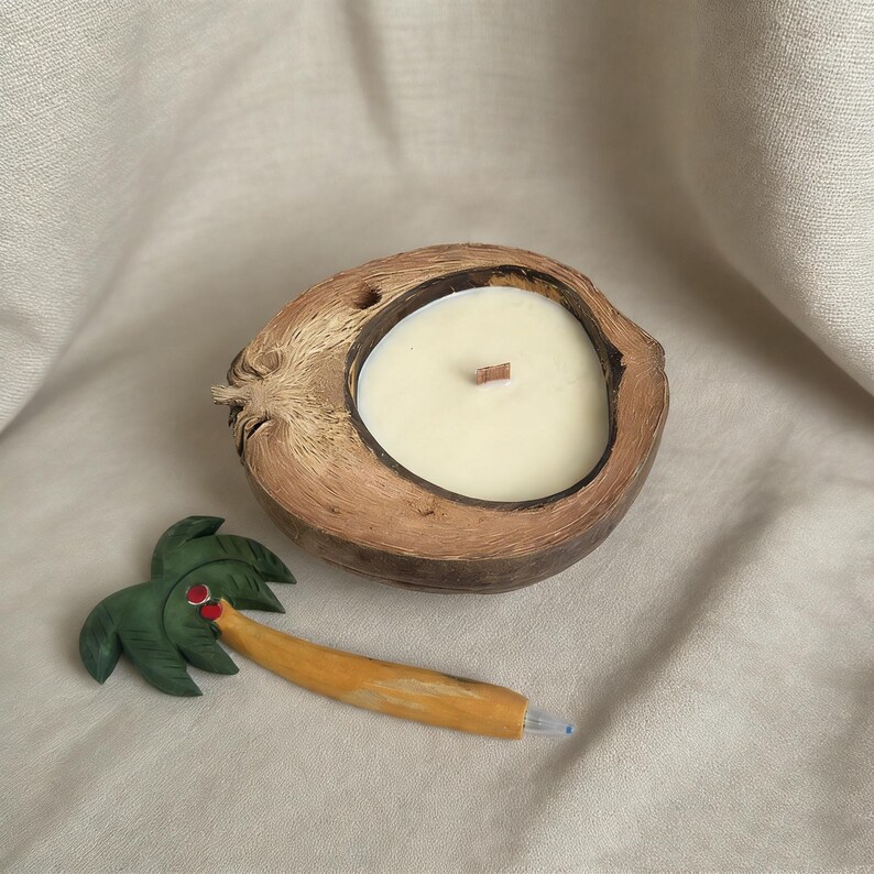 May include: A coconut shell candle with a wooden wick, filled with creamy wax. Beside it is a palm tree pen with green leaves, a red dot, and a yellow trunk. The candle and pen rest on a beige fabric.