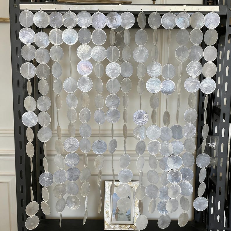 May include: A decorative curtain made of numerous, iridescent, round, shell-like discs strung together. The discs are primarily white with subtle hints of blue and gray. The curtain hangs from a dark metal frame, creating a shimmering, elegant effect.