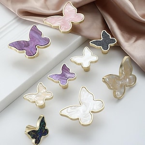 May include: A collection of butterfly-shaped cabinet knobs in various colors, including pink, purple, white, and black. Each knob features a gold-colored trim and a unique marbled or speckled pattern. These decorative knobs are ideal for adding a touch of elegance to furniture.