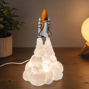 Könnte beinhalten: Eine Space-Shuttle-Lampe mit einem weiß-orangen Shuttle-Fahrzeug, das auf einer durchscheinenden, geometrischen weißen Wolkenbasis montiert ist, die ein sanftes Licht ausstrahlt. Das Shuttle hat schwarze Details an Flügeln und Rumpf.