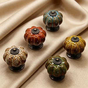 May include: A collection of five ceramic cabinet knobs in various colors, including blue, orange, yellow, green, and brown. Each knob has a pumpkin-like shape with a dark metal base and a decorative center. The knobs are arranged on a beige fabric.