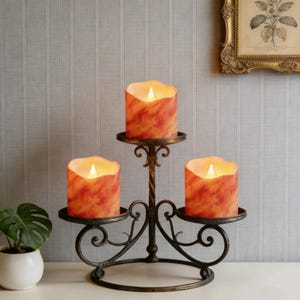 May include: A decorative candelabra holding three LED candles with an orange and red marbled effect. The dark metal candelabra has a twisted central column and scrolled arms. The candles have a warm, flickering glow, creating a cosy atmosphere.