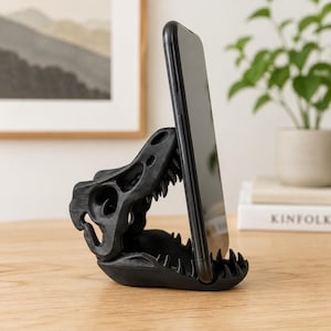 Könnte beinhalten: Ein mattschwarzer Smartphone-Ständer in Form eines Tyrannosaurus-Rex-Schädels. Das Gerät ruht im geöffneten Kiefer des Dinosauriers, wobei der Unterkiefer als Basis dient und die obere Schädelstruktur die Rückseite des Telefons stützt.