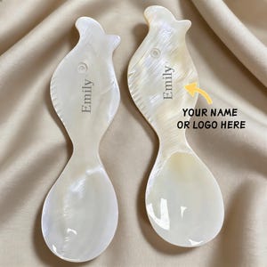 May include: Two pearlescent white gua sha tools with a unique, curved shape. Each tool has the name "Emily" engraved on it. The image also includes the text "YOUR NAME OR LOGO HERE" with an arrow, indicating customization options.