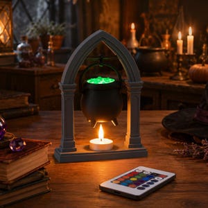 Può includere: Un calderone nero sospeso all'interno di una cornice grigia ad arco, con un liquido verde luminoso all'interno e una candela bianca sottostante. Un telecomando rettangolare con pulsanti colorati è posizionato in primo piano.