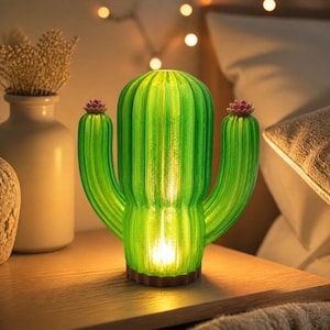 May include: A green cactus-shaped lamp with a ribbed texture and two arms, each topped with a small pink flower. The lamp emits a warm, glowing light from its base, illuminating the translucent green material of the cactus structure.