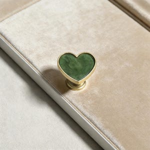 May include: A heart-shaped cabinet knob with a green, marbled center and a gold-colored metal frame. The knob is mounted on a beige surface, suggesting a furniture or decor item. The design is simple yet elegant.