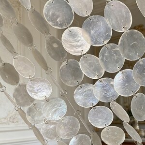 May include: A close-up of a decorative curtain made of iridescent, round and oval shell-like pieces. The pieces are strung together with silver-colored metal rings and chains, creating a shimmering, elegant effect. The curtain is likely used for interior decor.
