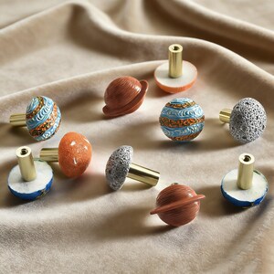 May include: A collection of decorative cabinet knobs with celestial designs. The knobs feature various planet and moon shapes in shades of blue, orange, brown, and gray, with gold-colored stems. The knobs are arranged on a soft, beige fabric.