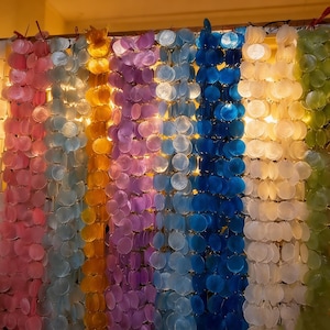 Może przedstawiać: Dekoracyjna zasłona wykonana z iryzujących, okrągłych kawałków muszli, ułożonych w pionowe paski. Zasłona prezentuje tęczę kolorów, w tym różowy, jasnoniebieski, żółty, fioletowy, ciemnoniebieski, biały i zielony. Muszle są połączone.