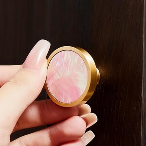May include: Circular cabinet knobs with gold-coloured rims. One knob has a pink and white swirl design, the other a colourful abalone shell pattern. They are mounted on a dark brown cabinet door.