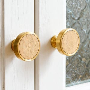 May include: Two cabinet knobs with a brass finish and a round, light wood center. The knobs are mounted on a white cabinet door with a glass panel featuring a decorative pattern. The knobs are a simple, yet elegant design.