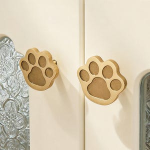 Op de afbeelding: Close-up van een kast met twee crèmekleurige deuren. Elke deur heeft een goudkleurige pootafdrukknop. De kastdeuren hebben een decoratief glaspaneel met een bloemmotief.