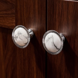 Può includere: Primo piano di due pomelli per mobili. Ogni pomello presenta un centro in marmo bianco con venature grigie, circondato da un bordo argentato e scanalato. I pomelli sono montati su un mobile in legno marrone scuro.