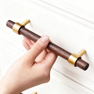 Pode incluir: Um puxador de armário de madeira castanha escura com detalhes dourados. A pega cilíndrica é segurada por uma mão contra um armário branco. O puxador tem cerca de 15 cm de comprimento.