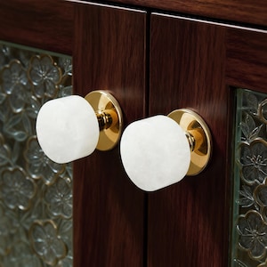 Può includere: Primo piano di due pomelli per ante di armadi. Ogni pomello presenta una pietra bianca e arrotondata su una base color oro. I pomelli sono montati su un'anta di armadio in legno marrone scuro. L'anta ha un pannello di vetro con un motivo floreale.