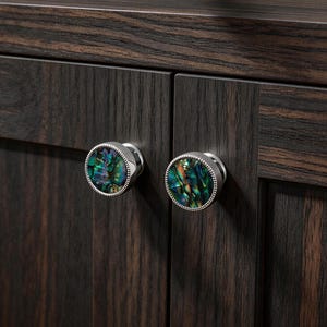 Peut inclure: Gros plan sur deux boutons de meuble avec des centres en coquille d'ormeau irisée et des bordures en métal argenté. Les boutons sont montés sur une armoire en bois brun foncé avec un grain de bois visible.