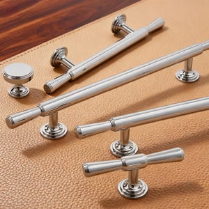 Pode incluir: Um conjunto de ferragens de armário prateadas polidas, incluindo puxadores e um botão. Os puxadores variam em tamanho e design, com uma barra cilíndrica e peças finais decorativas. O hardware é exibido em uma superfície texturizada bege.