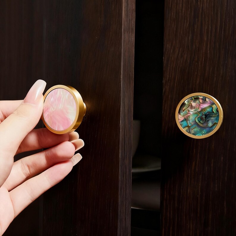 May include: Circular cabinet knobs with gold-coloured rims. One knob has a pink and white swirl design, the other a colourful abalone shell pattern. They are mounted on a dark brown cabinet door.