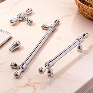 Può includere: Una collezione di ferramenta per mobili cromata lucida, tra cui maniglie e un pomello. Le maniglie hanno un design classico con estremità arrotondate e una barra centrale. Il pomello ha una forma piccola e arrotondata. La ferramenta è esposta su una superficie di marmo.