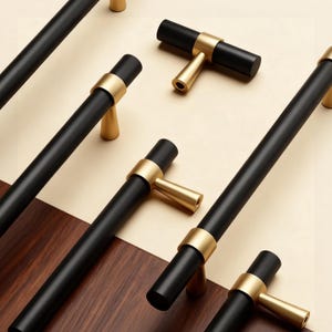 May include: A selection of black and gold cabinet pulls and handles. The pulls have a matte black finish with gold accents. The handles are T-shaped with a gold finish. The hardware is arranged on a wood and cream-coloured background.