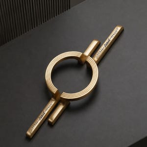 May include: A gold-toned metal door handle featuring a central circular ring with concentric grooved detailing. Two straight, flat bars extend from the ring, each engraved with the name Janet and a small leaf motif at the end of the handle.
