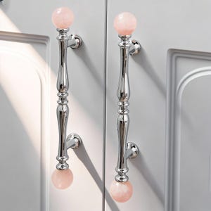 Op de afbeelding: Twee gepolijste chromen kastgrepen met een decoratief ontwerp. Elke handgreep heeft een roze, bolvormig kristal aan de boven- en onderkant. De handgrepen zijn gemonteerd op een witte kastdeur.