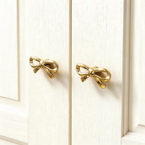 Pode incluir: Dois puxadores dourados em forma de laço montados numa porta de armário branca. Os puxadores são feitos de metal e têm um acabamento brilhante. A porta do armário tem painéis verticais.