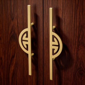 Op de afbeelding: Twee goudkleurige kastgrepen met een uniek ontwerp, gemonteerd op een donkere houten kast. Elke greep heeft een verticale staaf met een halfrond element. De kast heeft een rijke, donkerbruine houtnerf.