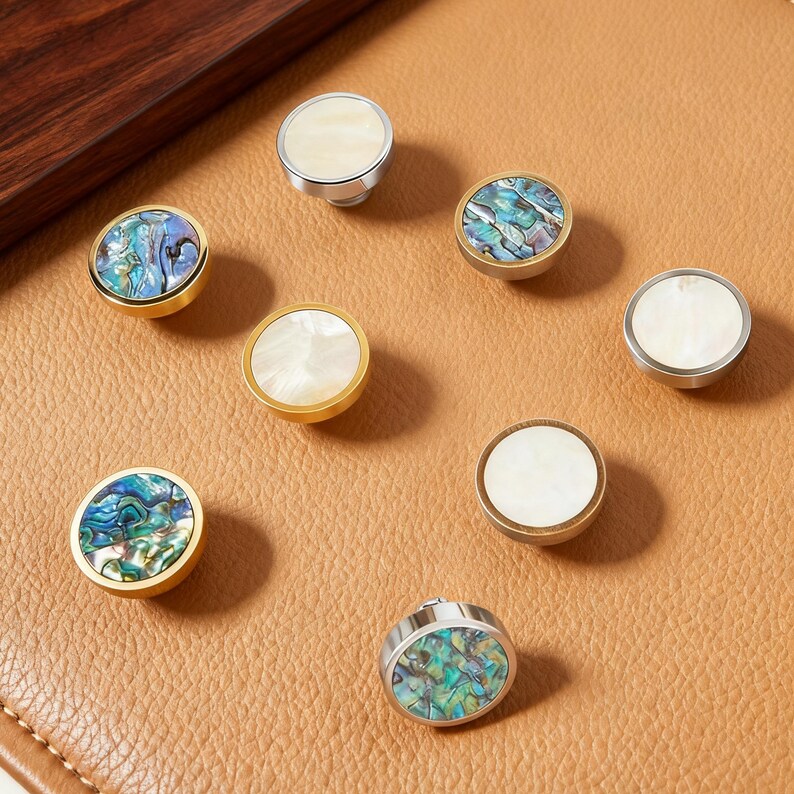 May include: A selection of decorative cabinet knobs with mother-of-pearl and abalone shell inlays. The knobs have gold and silver metal finishes. The knobs are round and have a flat top. The cabinet knobs are suitable for furniture.
