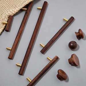 May include: A collection of wooden cabinet hardware, including long pulls and knobs. The pulls are dark brown with brass-colored accents. The knobs are various shapes and sizes, all in a matching dark wood finish.