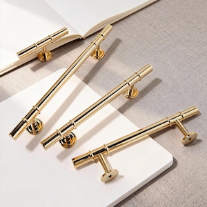 May include: Four gold-colored cabinet pulls with a cylindrical design. The pulls are arranged on a white surface, with a book in the background. The pulls have a T-bar shape and are made of a shiny, metallic material.