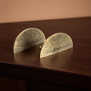 May include: Two semi-circular brass drawer pulls featuring a sunburst texture and engraved with the name Jones in a cursive script. Each piece has a metallic finish with dark etched lines radiating from the center toward the curved edge.