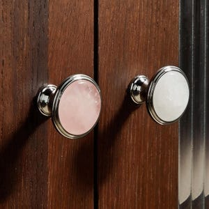May include: Two cabinet knobs with a polished silver finish. One knob features a pink stone center, the other a white stone center. Both are set against a dark wood cabinet door.