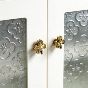 Op de afbeelding: Close-up van een witte kastdeur met twee messingkleurige klavervormige knoppen. De kastdeur heeft een glazen paneel met een bloemmotief. De knoppen zijn gedetailleerd met bladnerven.