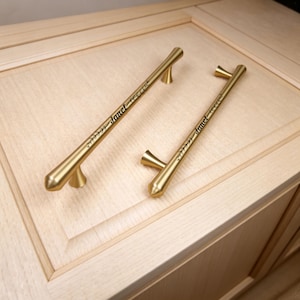 May include: Two brushed gold metal cabinet pulls with tapered ends and conical mounting posts. Each handle features a laser-engraved design consisting of a small leaf branch motif and the name Janet in a cursive script font centered on the bar.