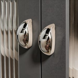 Puede incluir: Primer plano de dos tiradores de armario cromados pulidos con diseño de lágrima. Los tiradores están montados en una puerta de armario gris oscuro. El acabado cromado refleja el entorno, creando un efecto espejo. La puerta del armario es de color mate sólido.