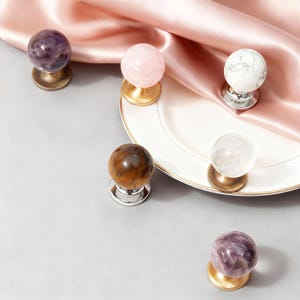 Puede incluir: Colección de tiradores decorativos para armarios con tapas esféricas de piedras preciosas. Los tiradores presentan amatista, cuarzo rosa, mármol, cuarzo transparente y ojo de tigre. Cada tirador está montado sobre una base metálica.