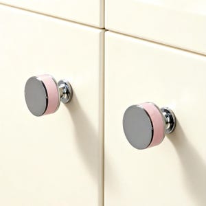 May include: Two cabinet knobs with a silver disc face and a pink band. The knobs are mounted on a cream-coloured cabinet door. The knobs are a stylish addition to any cabinet.