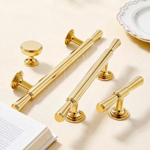 May include: A collection of gold-colored cabinet hardware, including two long bar pulls, two smaller pulls, and a round knob. The hardware has a shiny finish and a classic design, suitable for kitchen or bathroom cabinets.