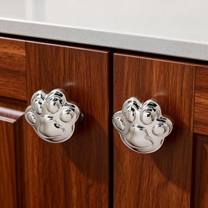 May include: Two polished chrome paw print cabinet knobs mounted on a dark wood cabinet. The knobs are shaped like a dog or cat paw, with five circular pads and a smooth, reflective surface. The cabinet has a rich, reddish-brown finish.