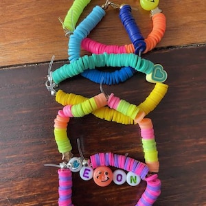 May include: A collection of colorful beaded bracelets in various shades of blue, pink, green, yellow, and orange. Some bracelets feature smiley face and heart charms, and one spells out the name "EON" with letter beads.