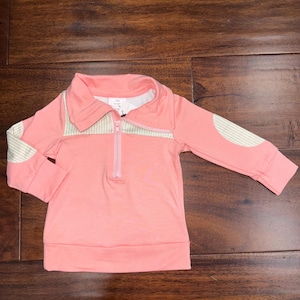 May include: A light pink long-sleeved shirt with a zip-up collar. The shirt features a light green and white striped design on the collar and elbow patches. The shirt is made of a soft material.