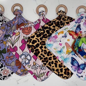 May include: A collection of baby security blankets with wooden ring attachments. The blankets feature various patterns, including bows, cow print, floral designs, leopard print, butterflies, and Disney princess illustrations. Each blanket has a square shape.