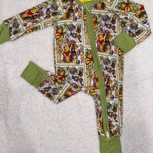May include: A baby's one-piece pajama with a colorful cartoon print featuring Winnie the Pooh and friends. The pajamas have long sleeves and legs with green cuffs and trim, and a full-length zipper. A yellow tag with text is attached.