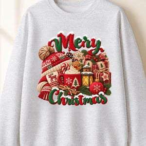 May include: A light grey crewneck sweatshirt featuring a festive graphic print with the text "Merry Christmas". The design includes a red and white knit hat, a red mug with a gingerbread man, wrapped gift boxes, and a glowing lantern.