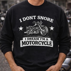 Może przedstawiać: Czarna bluza z napisem "I DON'T SNORE" nad grafiką motocykla i napisem "I DREAM I'M A MOTORCYCLE". Projekt zawiera dwie gwiazdki. Bluza ma okrągły dekolt.