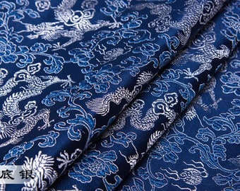 May include: Close-up of a folded, dark blue fabric with a silver dragon and floral pattern. The fabric has a shiny, textured appearance. The Chinese characters in the image translate to "Tibetan blue silver".