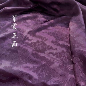 May include: A deep purple fabric featuring a tonal jacquard pattern of stylized clouds. The material has a smooth, lustrous finish with visible folds and drapes. The Chinese characters 紫棠正面 are printed vertically in white on the left side.